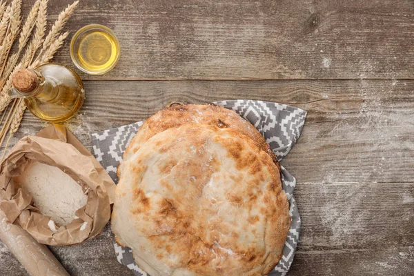 Top view of lavash bread on gray towel with pattern near wheat spikes, rolling pin, flour package and bottle of olive oil on wooden table — Stock Photo