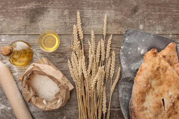 Vista dall'alto del pane lavash su asciugamano grigio vicino a punte di grano, mattarello, pacchetto di farina aperta e bottiglia di olio d'oliva su tavolo di legno — Foto stock