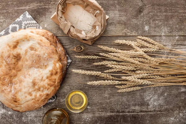 Top view of lavash bread on towel with pattern near wheat spikes, rolling pin, flour and olive oil on wooden table — Stock Photo