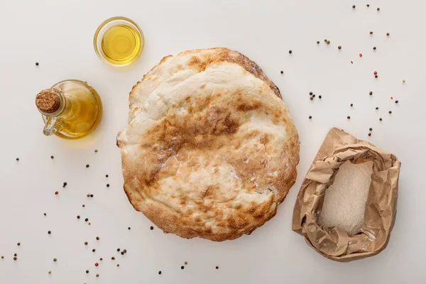 Vista superior del pan de lavash cerca de harina y aceite de oliva en la superficie blanca con granos de pimienta - foto de stock