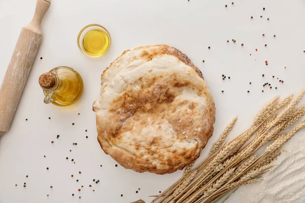 Vista superior del pan de lavash cerca de espigas y aceite de oliva en la superficie blanca con granos de pimienta - foto de stock