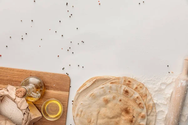 Vue du dessus du pain de lavande plat près de la planche à découper en bois avec farine et huile d'olive sur surface blanche avec grains de poivre — Photo de stock