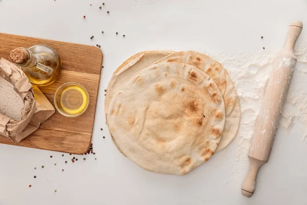 Vista dall'alto del pane lavash piatto vicino al mattarello e tagliere in legno con farina e olio d'oliva su superficie bianca con grani di pepe — Foto stock