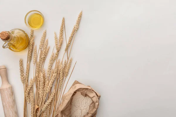 Vista dall'alto di punte di grano, mattarello, olio d'oliva e pacchetto di farina sulla superficie bianca — Foto stock