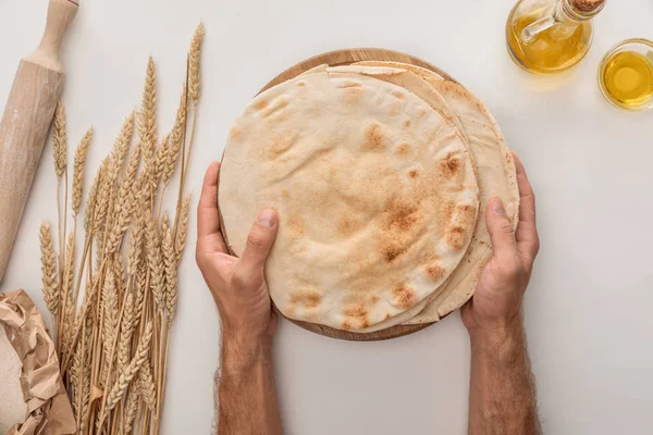 Vista ritagliata dell'uomo che tiene piatto pane lavash vicino a punte di grano, mattarello e olio d'oliva sulla superficie bianca — Foto stock