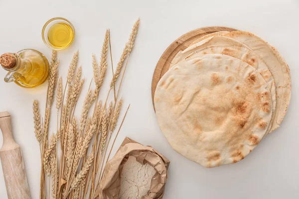 Vista dall'alto di pane lavash piatto vicino a punte di grano, mattarello e olio d'oliva su superficie bianca — Foto stock