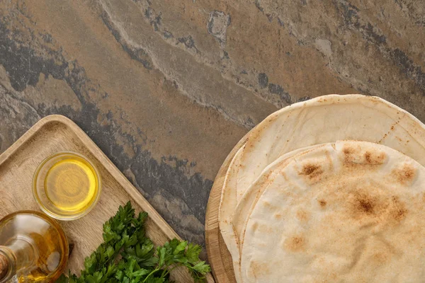 Vista dall'alto del pane lavash piatto vicino a piatto di legno con prezzemolo e olio d'oliva su superficie di marmo marrone — Foto stock