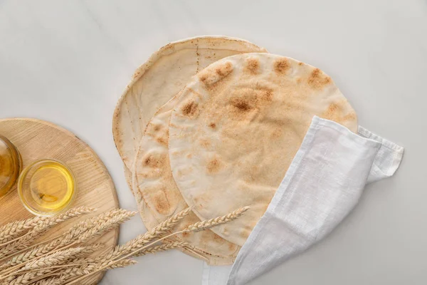 Vue du dessus du pain lavash plat recouvert d'une serviette blanche près de la planche à découper avec des pointes et de l'huile sur la surface du marbre — Photo de stock