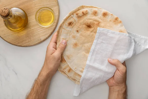 Cropped view of man holding flat lavash bread covered with white towel near cutting board oil on marble surface — Stock Photo