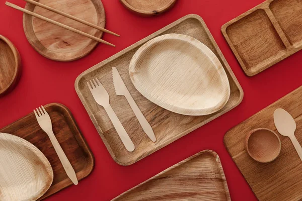 Vista superior de platos de madera ecológicos, platos, palillos y cubiertos sobre fondo rojo - foto de stock