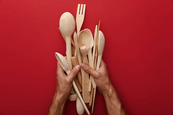 Vista recortada del hombre sosteniendo cucharas de madera, tenedor y utensilios de cocina sobre fondo rojo - foto de stock