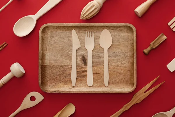 Vista dall'alto di piatto di legno rettangolare naturale con posate su sfondo rosso con utensili da cucina — Foto stock