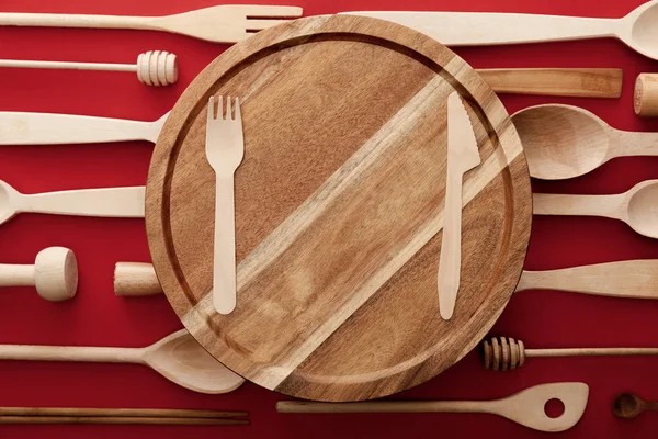 Vista superior da placa de corte de madeira redonda com faca e garfo no fundo vermelho com utensílios de cozinha — Fotografia de Stock