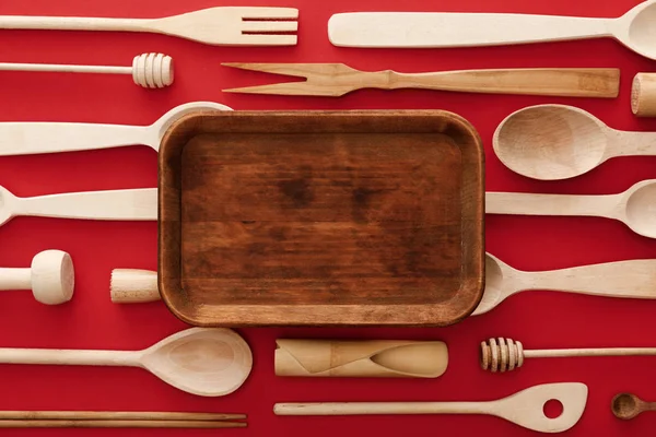Vue du dessus du plat rectangulaire vide en bois sur fond rouge avec ustensiles de cuisine — Photo de stock