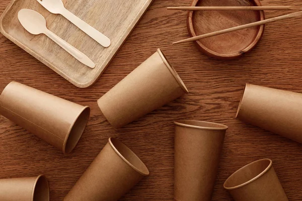 Vista superior de platos de madera, cubiertos, palillos y vasos de papel sobre fondo marrón - foto de stock