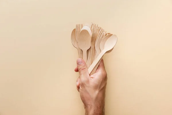 Cropped view of man holding natural wooden spoons and forks on beige background — Stock Photo