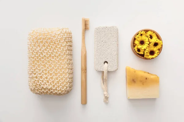 Acostado plano con esponja de baño cerca del cepillo de dientes, pedazo de jabón, piedra pómez y taza con flores sobre fondo blanco - foto de stock