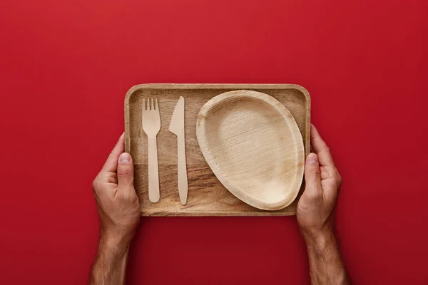 Cropped view of man holding natural rectangular wooden dish with plate, fork and knife on red background — Stock Photo