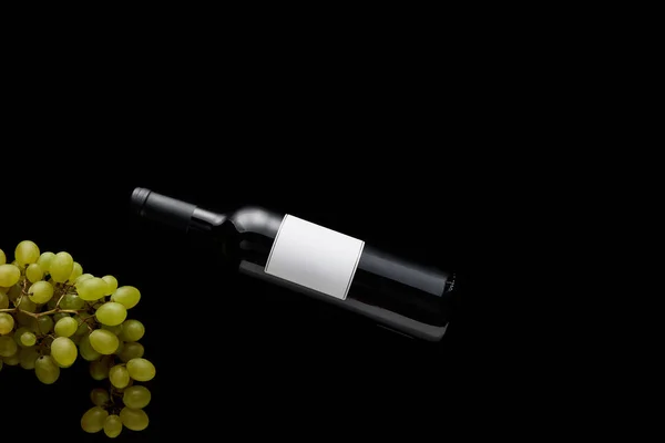 Вид зверху пляшки вина з порожньою білою етикеткою біля стиглого винограду ізольовано на чорному — стокове фото