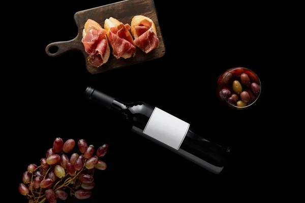 Вид зверху пляшки вина з порожньою білою етикеткою біля стиглого винограду, оливок і нарізаного прошутто на багеті ізольовані на чорному — стокове фото