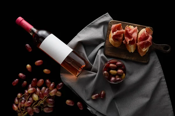Vue du dessus de la bouteille de vin avec étiquette blanche vierge sur une serviette grise près du raisin mûr, des olives et du prosciutto tranché sur une baguette isolée sur du noir — Photo de stock