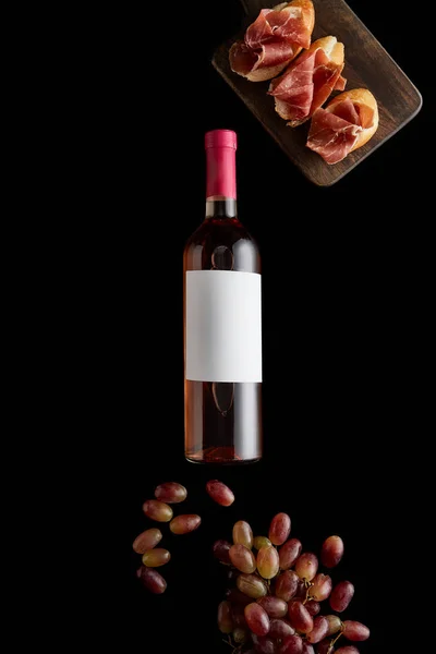Top view of rose wine bottle with blank white label near ripe grape and sliced prosciutto on baguette isolated on black — Stock Photo
