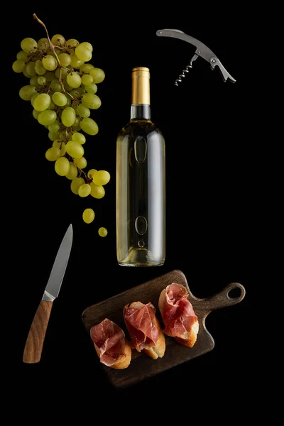 Top view of bottle with white wine near grape, knife, corkscrew and sliced prosciutto on baguette isolated on black — Stock Photo