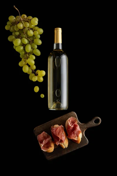 Vista dall'alto di bottiglia con vino bianco vicino all'uva e prosciutto affettato su baguette isolate su nero — Foto stock