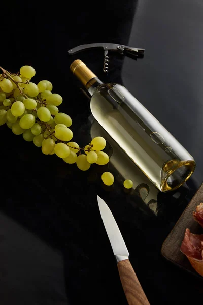 Bottle with white wine near grape, knife and corkscrew on black surface — Stock Photo
