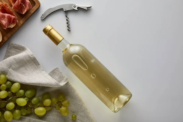 Top view of bottle with white wine near grape, corkscrew and sliced prosciutto on baguette on white background — Stock Photo