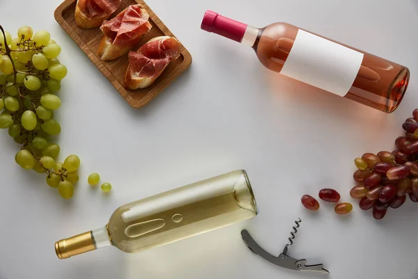 Vista superior de garrafas com vinho branco e rosa perto de uva, saca-rolhas e prosciutto fatiado na baguete no fundo branco — Fotografia de Stock