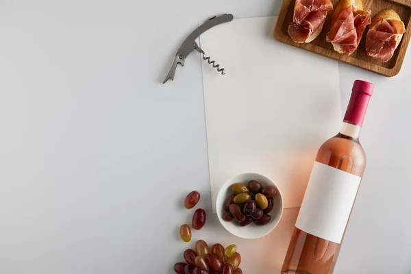 Vista superior de la botella con vino rosa cerca de la uva, sacacorchos, aceitunas, papel en blanco y jamón en rodajas en baguette sobre fondo blanco - foto de stock