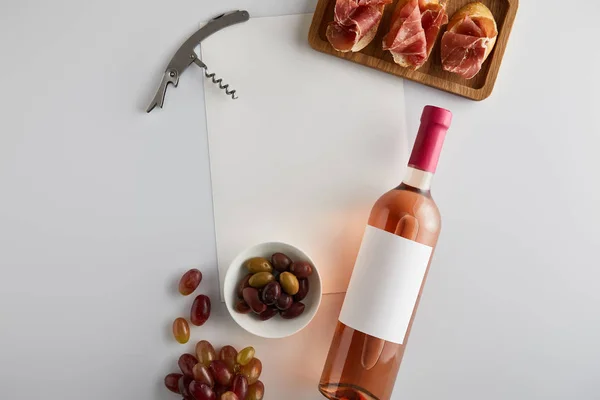 Vue du dessus de la bouteille avec vin rose près du raisin, tire-bouchon, olives, papier blanc et prosciutto tranché sur baguette sur fond blanc — Photo de stock
