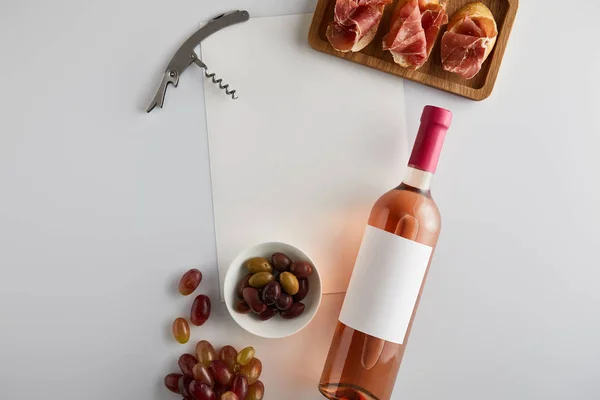 Vista superior de la botella con vino rosa cerca de la uva, sacacorchos, aceitunas, papel en blanco y jamón en rodajas en baguette sobre fondo blanco - foto de stock