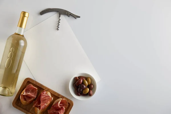 Vue du dessus de la bouteille avec vin blanc près des olives, tire-bouchon, papier blanc et prosciutto tranché sur baguette sur fond blanc — Photo de stock