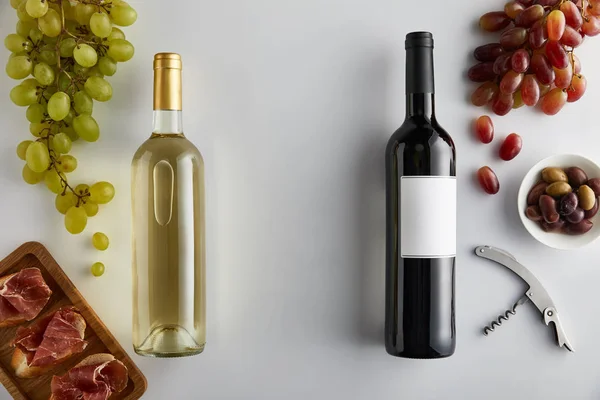 Top view of bottles with white and red wine near grape, corkscrew, olives and sliced prosciutto on baguette on white background — Stock Photo