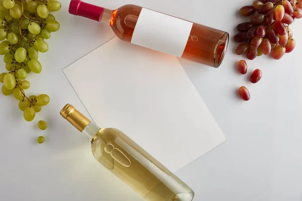 Vista superior de botellas con vino blanco y rosa cerca de la uva y papel en blanco sobre fondo blanco - foto de stock