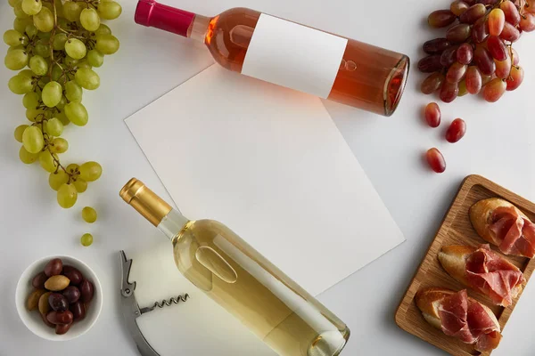 Top view of bottles with white and rose wine near blank paper, grape, corkscrew, olives and sliced prosciutto on baguette on white background — Stock Photo