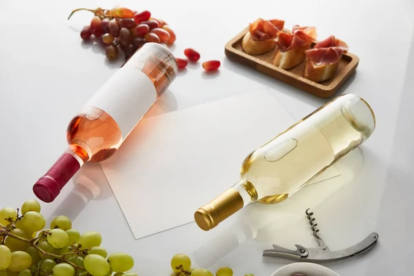 Garrafas com branco, vinho rosa perto de uva, papel em branco, saca-rolhas e prosciutto fatiado na baguete em fundo branco — Fotografia de Stock