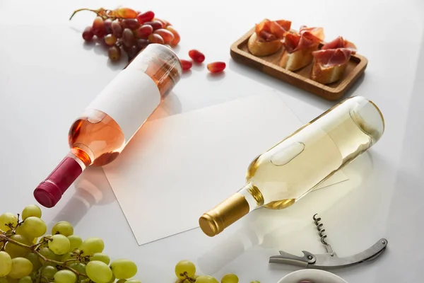 Bottles with white, rose wine near grape, blank paper, corkscrew and sliced prosciutto on baguette on white background — Stock Photo