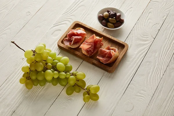 Trauben, Prosciutto auf Baguette, Oliven auf weißer Holzoberfläche — Stockfoto