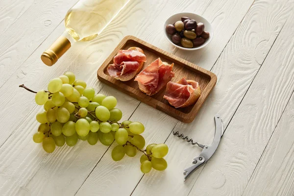 Flasche mit Weißwein in Traubennähe, Schinken auf Baguette, Oliven und Korkenzieher auf weißer Holzoberfläche — Stockfoto