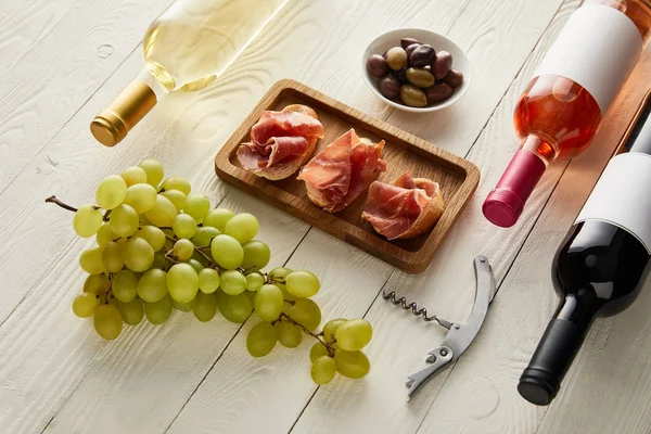Бутылки с красным, белым и розовым вином рядом с виноградом, прошутто на багете возле оливок и штопор на белой деревянной поверхности — стоковое фото