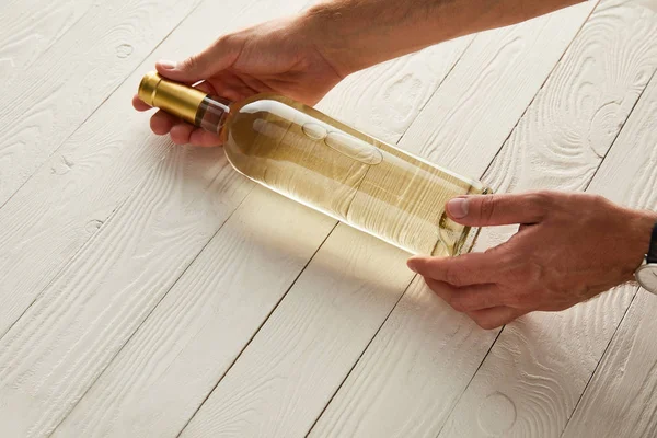 Cropped view of man holding bottle of white wine on white wooden surface — Stock Photo