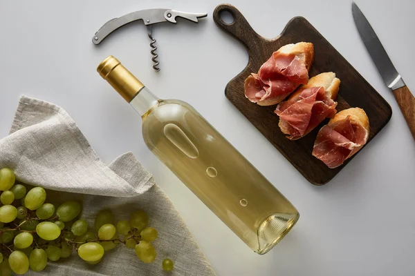 Top view of bottle with white wine near grape, corkscrew, knife and sliced prosciutto on baguette on white background — Stock Photo