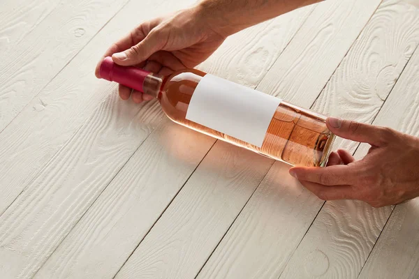 Vista cortada do homem segurando garrafa de vinho rosa com rótulo em branco na superfície de madeira branca — Fotografia de Stock