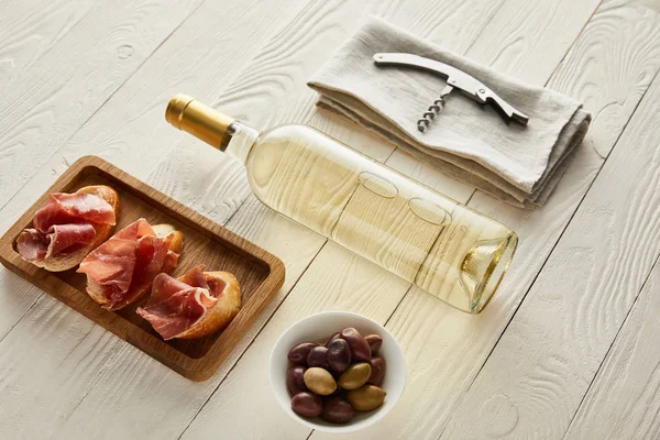 Bottle with white wine near prosciutto on baguette, olives and corkscrew on napkin on white wooden surface — Stock Photo