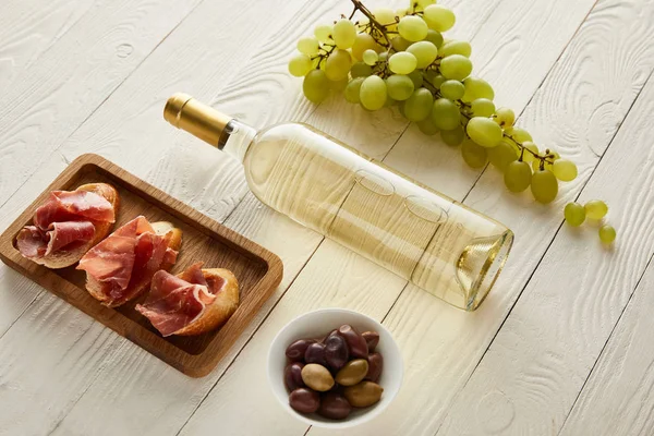 Botella con vino blanco cerca de la uva, jamón en baguette, aceitunas en la superficie de madera blanca - foto de stock