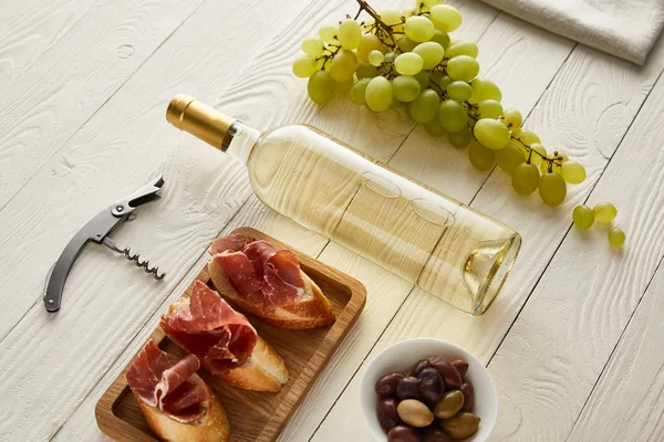 Bottle with white wine near grape, prosciutto on baguette, olives and corkscrew on white wooden surface — Stock Photo