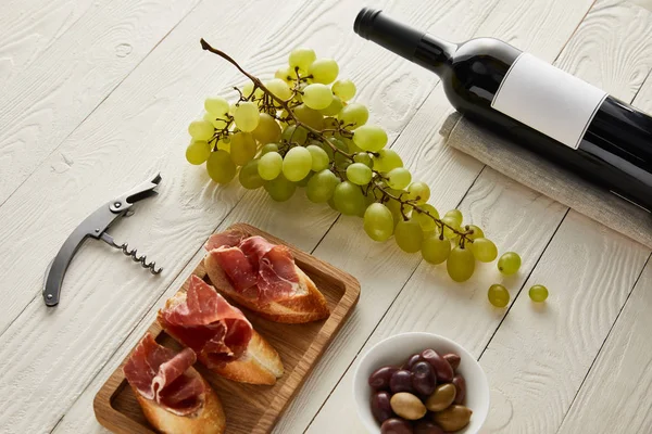 Flasche mit Wein in Traubennähe, Schinken auf Baguette, Oliven und Korkenzieher auf weißer Holzoberfläche — Stockfoto